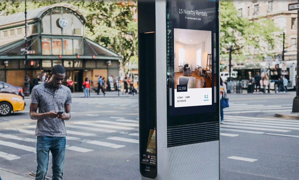 New York City kiosk with a digital ad on the side, a man looking at his cell phone standing to the left of the kiosk, city in the background.
